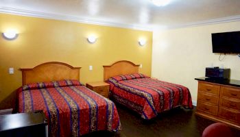 A hotel room features two double beds with striped bedding, a wall-mounted TV, dresser, nightstand, and warmly lit walls.