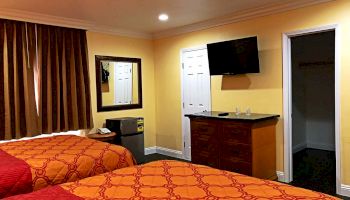 The image shows a hotel room with two beds and patterned bedspreads. There's a wall-mounted TV, a mirror, a small fridge, and a doorway to a bathroom.