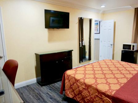 The image shows a cozy, simply furnished hotel room with a bed, TV, dresser, chair, microwave, and a small fridge beneath a window with brown curtains.