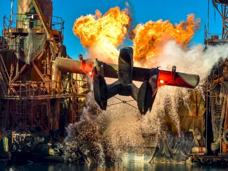 A seaplane splashes into water amidst an industrial setting with explosions and fire in the background, creating a dramatic and action-packed scene.